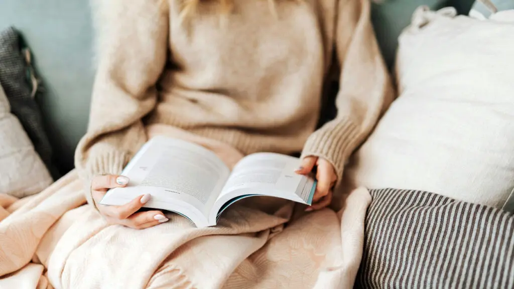 A person sits under a blanket with a book in their lap and their face is not seen.