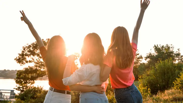 Three women with the backs to the camera face the sun and raise their arms up.