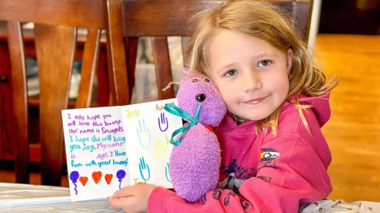 A little girl holds a purple stuffed animal and a letter to a senior citizen.