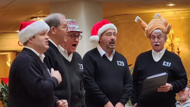 A group of five men are singing. Two are in Santa hats and one is in a turkey hat.