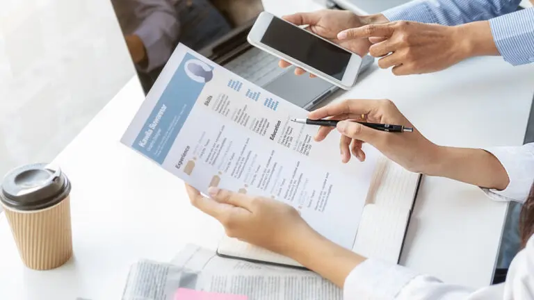 One pair of hands hold a resume over newspaper now hiring ads and another hand hold a cell phone over a laptop.