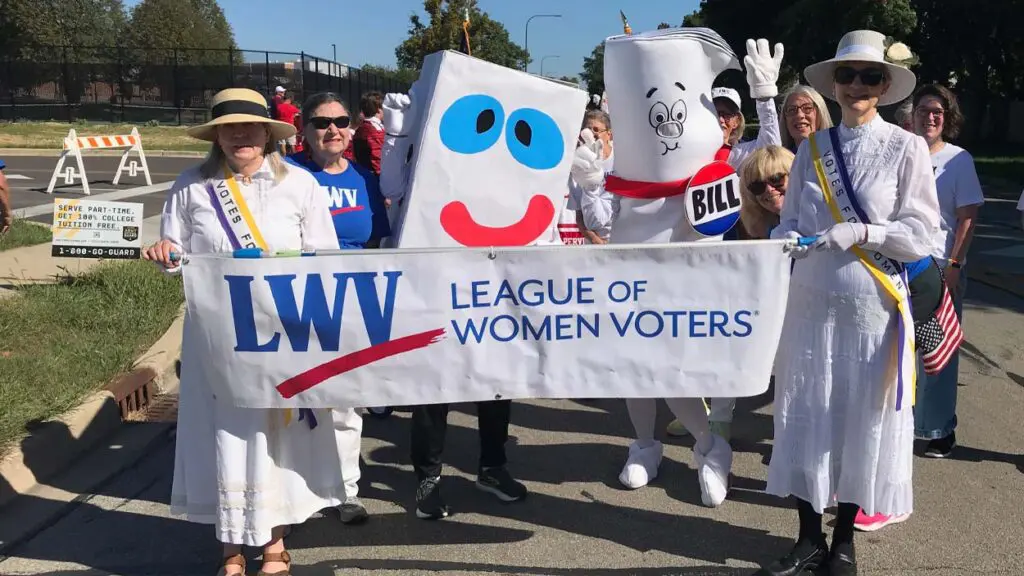 Women dressed as suffragettes hold a League of Women Voters of Naperville banner while walking in a parade. Others are dressed as giant ballot boxes.