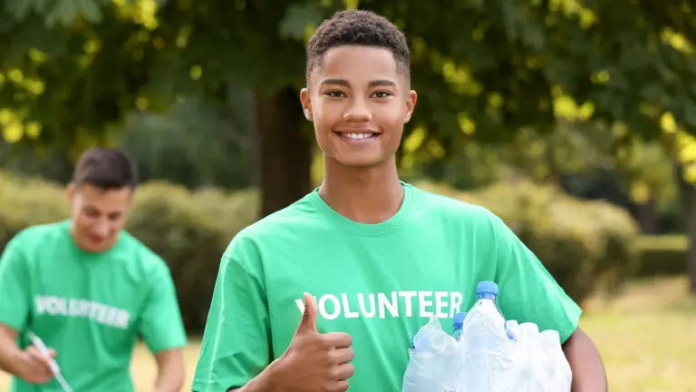 A young man smiles that the camera with his right hand giving a thumbs up. He's wearing a green shirt that says "Volunteer"