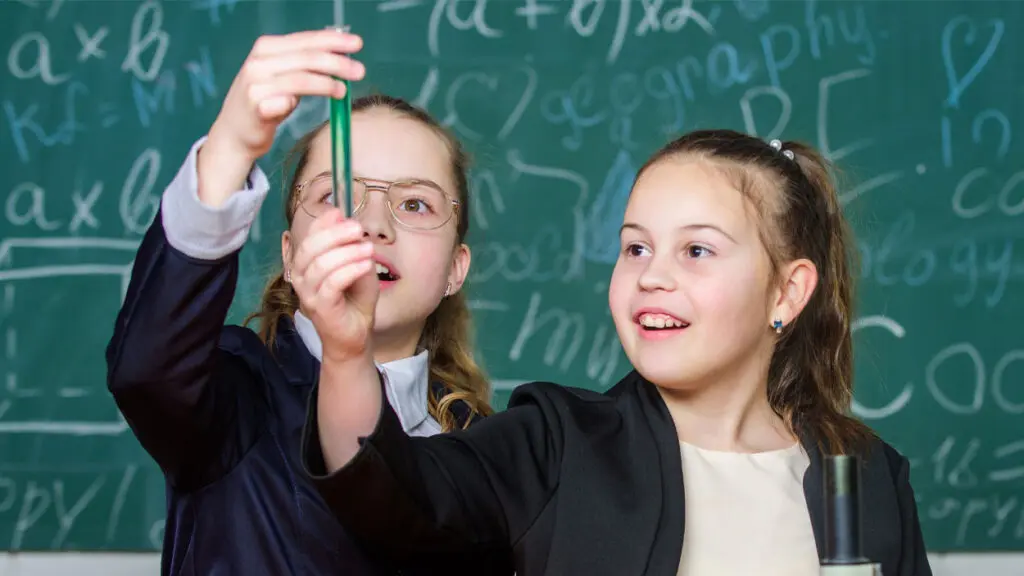 two young girls stand in front of a green chalkboard. They hold up a test tube filled with a green substance.