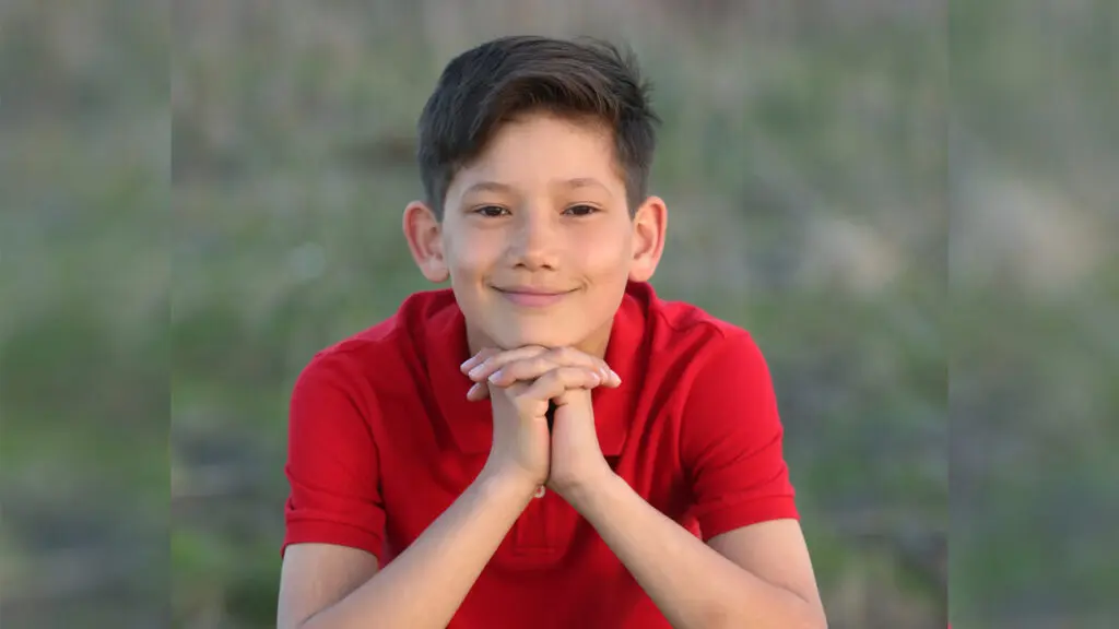 A young boy (He is Max of Max's Mission). He is dressed in a red shirt and rests his chin on his clasped hands and softly smiles.