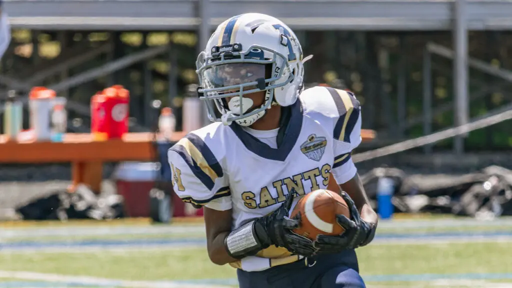 A boy is on a football field in Naperville Saints football gear holding a football.