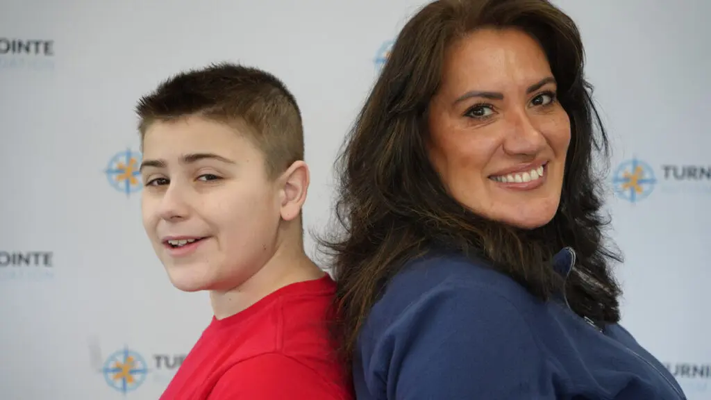 A young boy in a red t-shirt stands with his back to a woman in a blue shirt. They are in front of a background that says turning pointe autism foundation.