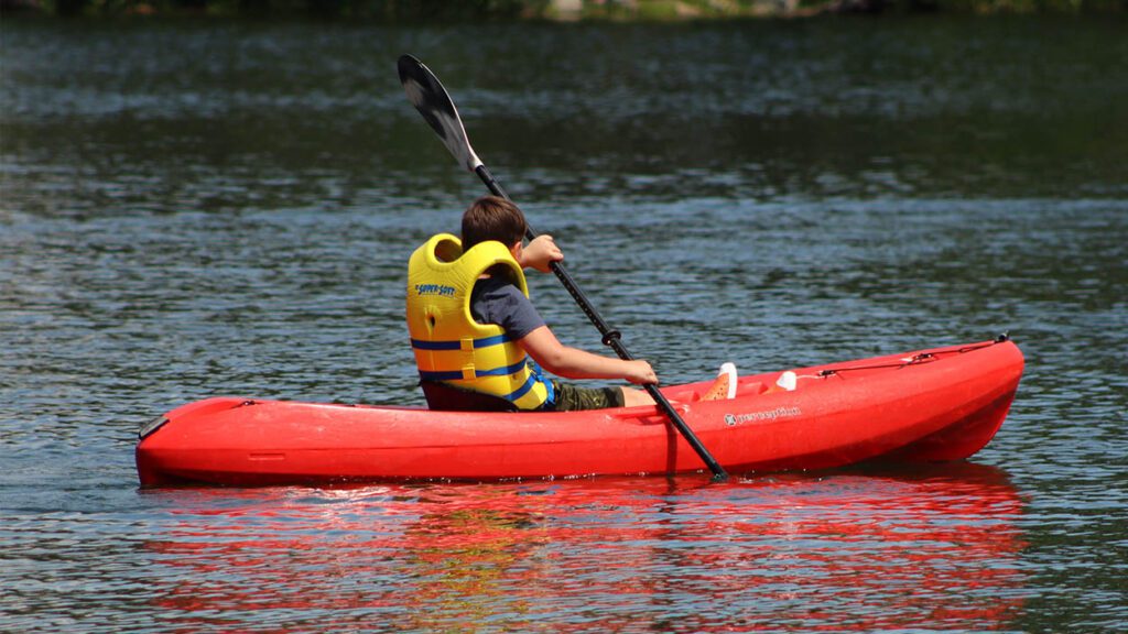 A young boy in a yellow life jackets rows a canoe on the water