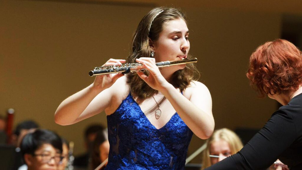 A young woman in a blue dress plays the flute.