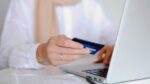 Close up of a woman's hand holding a credit card with one hand and navigating on a laptop with the other.