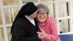 A nun hugs and kisses an older woman in a pink sweater who smiles